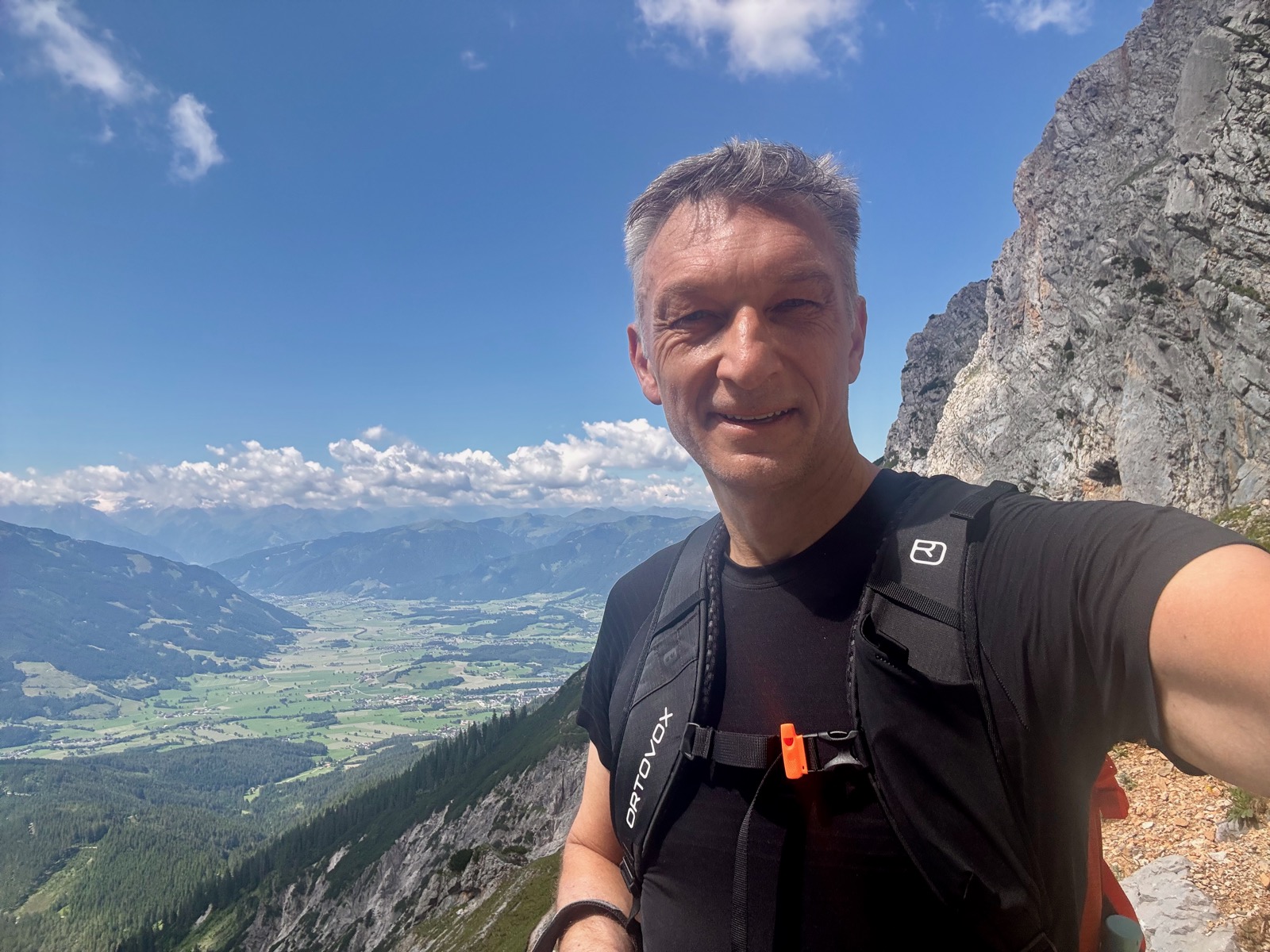 A hiker on a narrow rock ledge high above a green valley, a limestone face rising alongside