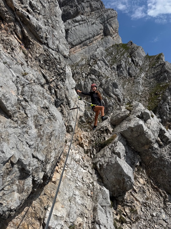A climber in helmet and harness, clipped to the fixed cable on a steep limestone face