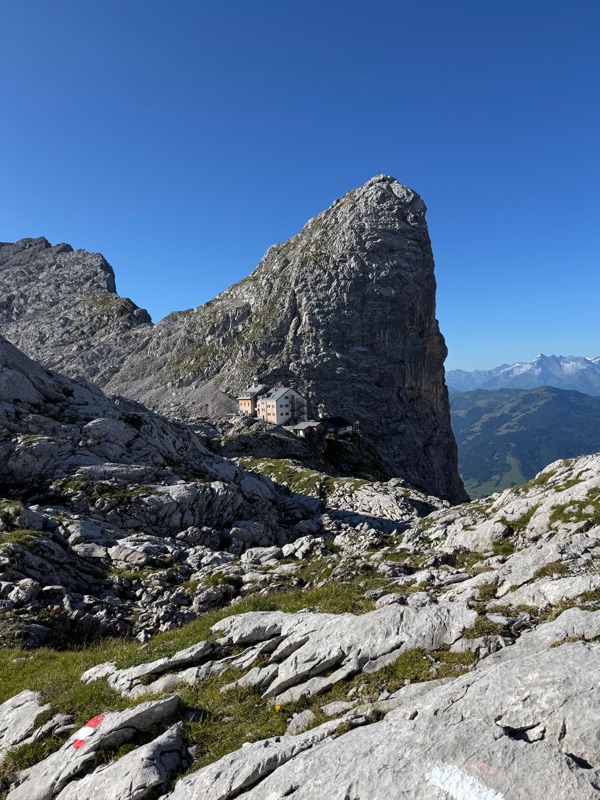 An alpine hut dwarfed by a huge limestone tower, the valley far below