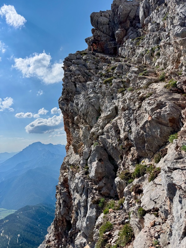An exposed traverse along a cliff edge, with a long drop to a green valley and distant peaks
